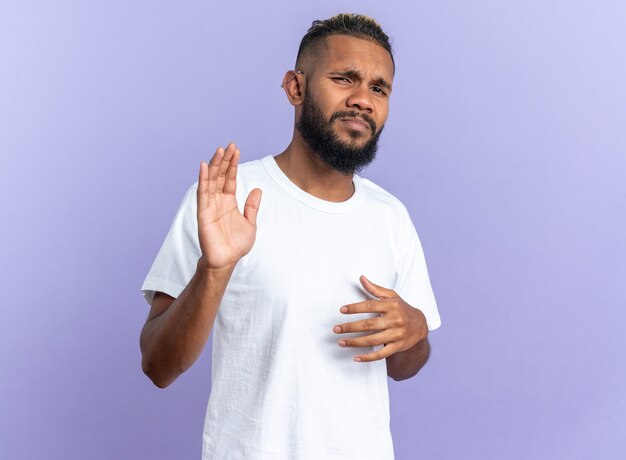 Joven afroamericano en camiseta blanca mirando a cámara confundido y disgustado haciendo gesto de parada con las manos de pie sobre fondo azul.