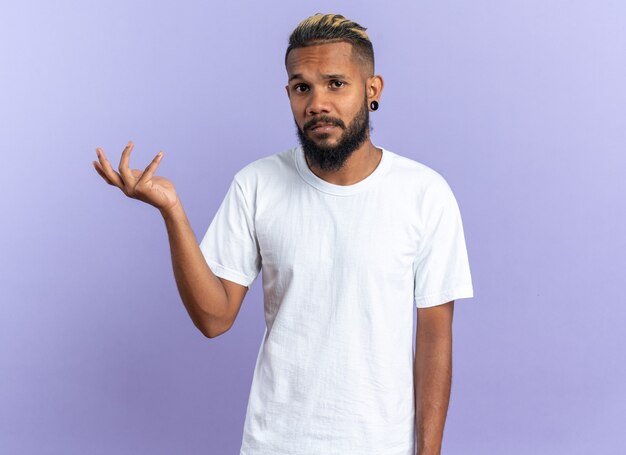 Joven afroamericano en camiseta blanca mirando a la cámara con expresión confusa levantando el brazo con indignación de pie sobre fondo azul.