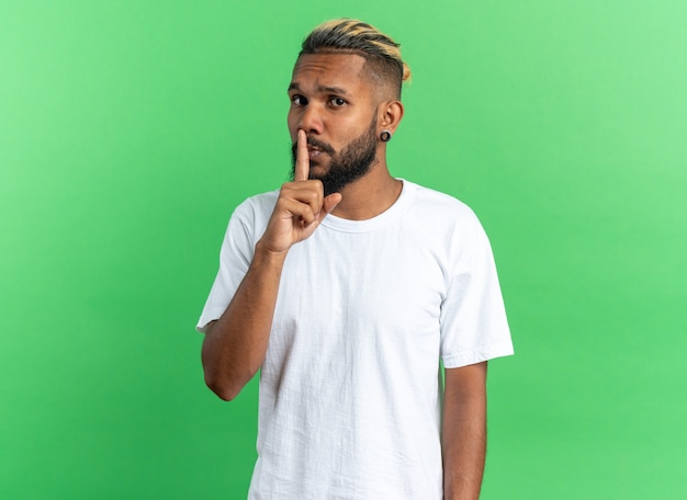 Joven afroamericano en camiseta blanca mirando a cámara con rostro serio haciendo gesto de silencio con el dedo en los labios de pie sobre fondo verde