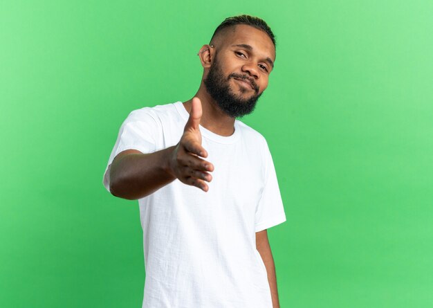 Joven afroamericano en camiseta blanca mirando a la cámara sonriendo amable ofreciendo saludo de mano de pie sobre fondo verde