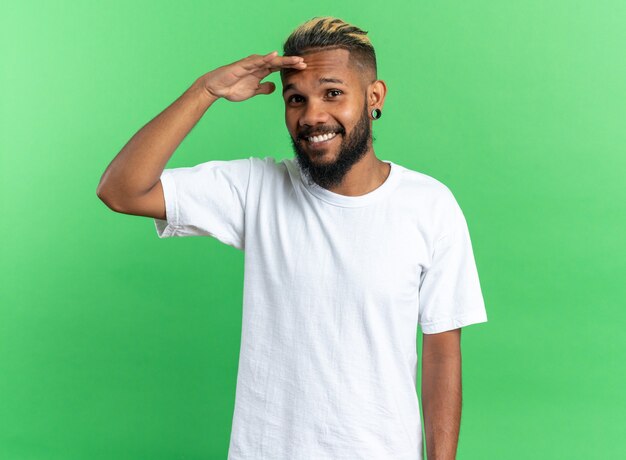 Joven afroamericano en camiseta blanca mirando a la cámara sonriendo con la mano en la frente de pie sobre fondo verde