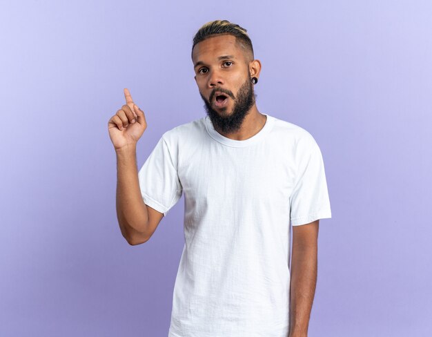 Joven afroamericano en camiseta blanca mirando a cámara sorprendido mostrando el dedo índice con nueva gran idea de pie sobre fondo azul.