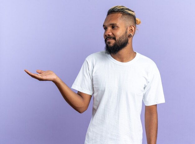 Joven afroamericano en camiseta blanca mirando a un lado con el brazo hacia fuera presentando copia espacio sonriente de pie sobre fondo azul.