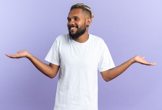 Joven afroamericano en camiseta blanca mirando a un lado sonriendo alegremente extendiendo los brazos a los lados de pie sobre fondo azul.