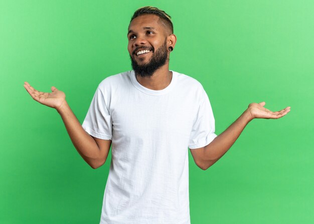 Joven afroamericano en camiseta blanca mirando a un lado sonriendo ampliamente extendiendo los brazos a los lados de pie sobre fondo verde