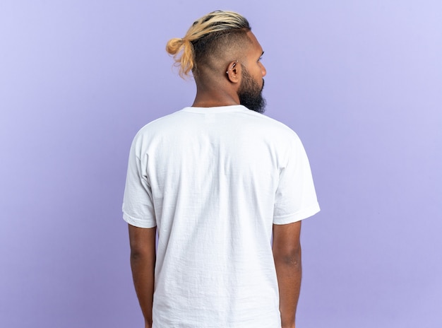 Joven afroamericano en camiseta blanca de pie con la espalda sobre fondo azul.