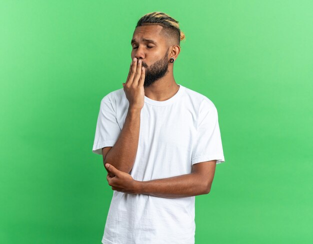 Joven afroamericano en camiseta blanca que sopla un beso feliz y seguro de pie sobre fondo verde