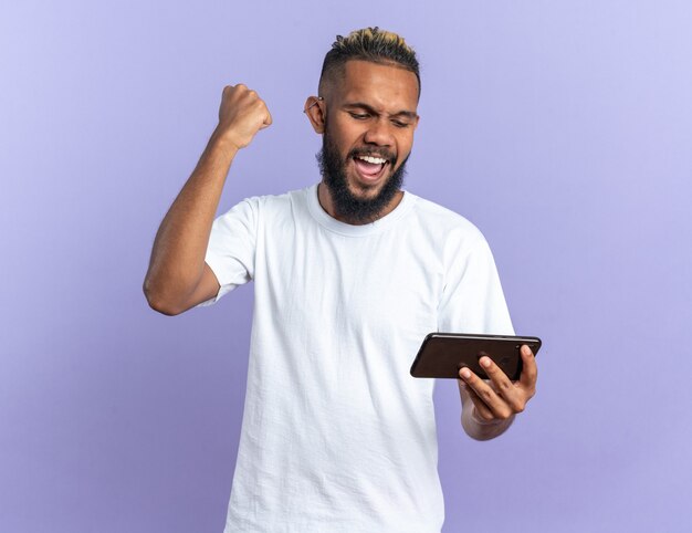 Joven afroamericano en camiseta blanca con smartphone apretando el puño feliz y emocionado gritando regocijándose por su éxito de pie sobre fondo azul.