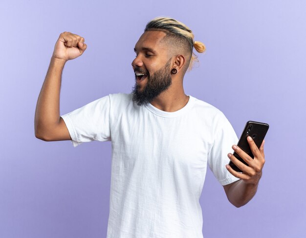 Joven afroamericano en camiseta blanca con smartphone apretando el puño feliz y emocionado gritando regocijándose por su éxito