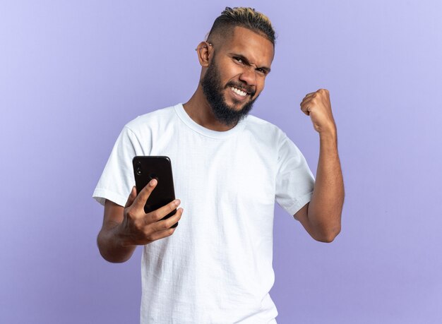 Joven afroamericano en camiseta blanca con smartphone apretando el puño feliz y emocionado regocijándose de su éxito de pie sobre fondo azul.