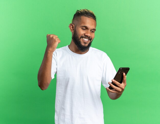 Joven afroamericano en camiseta blanca con smartphone apretando el puño feliz y emocionado regocijándose de su éxito