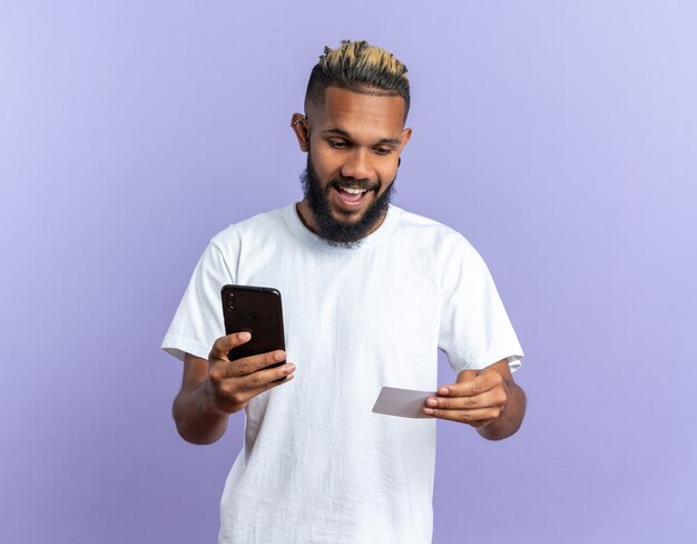 Joven afroamericano en camiseta blanca sosteniendo smartphone mirando su tarjeta de crédito feliz y sorprendido de pie sobre fondo azul.