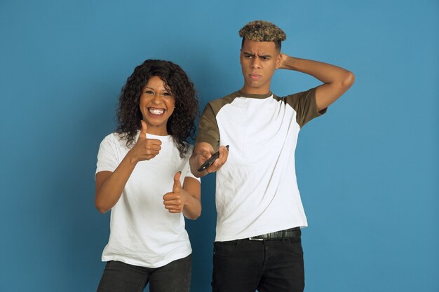 Joven afroamericano emocional y mujer posando sobre fondo azul. Hermosa pareja. Concepto de emociones humanas, expresión facial, relaciones, anuncio. Vean la televisión juntos, su canal favorito.