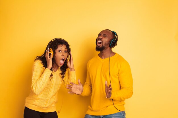 Joven afroamericano emocional y mujer en ropa casual brillante posando sobre fondo amarillo. Hermosa pareja. Concepto de emociones humanas, expresión facial, anuncio. Sorprendido escuchar música.