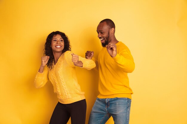 Joven afroamericano emocional y mujer en ropa casual brillante sobre fondo amarillo. Hermosa pareja. Concepto de emociones humanas, expresión facial, relaciones, anuncio. Bailando y cantando.