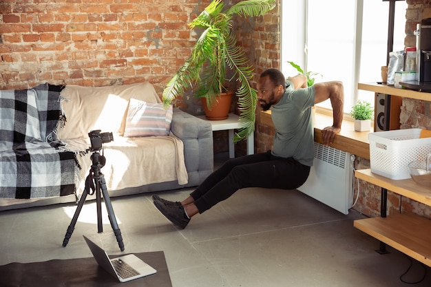 Joven afroamericano enseñando en casa cursos en línea de acondicionamiento físico, estilo de vida aeróbico y deportivo durante la cuarentena, grabando en cámara, transmitiendo