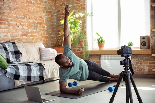 Joven afroamericano enseñando en casa cursos en línea de acondicionamiento físico, estilo de vida aeróbico y deportivo durante la cuarentena, grabando en cámara, transmitiendo