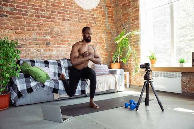 Foto gratuita joven afroamericano enseñando en casa cursos en línea de acondicionamiento físico, estilo de vida aeróbico y deportivo durante la cuarentena, grabando en cámara, transmitiendo