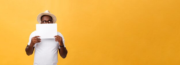 Joven afroamericano feliz escondido detrás de un papel en blanco aislado de fondo amarillo