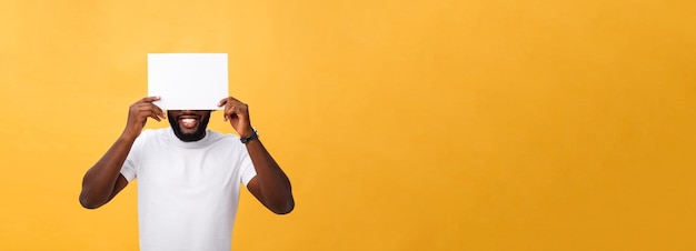 Foto gratuita joven afroamericano feliz escondido detrás de un papel en blanco aislado de fondo amarillo