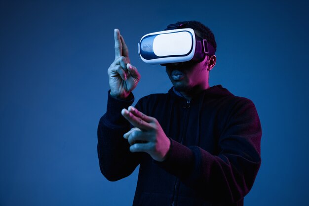 Joven afroamericano en gafas VR en neón sobre fondo azul. Retrato masculino