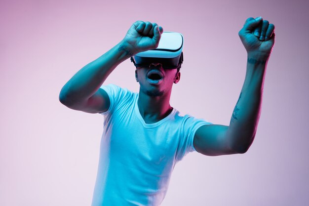 Joven afroamericano jugando y usando gafas VR en luz de neón sobre fondo degradado