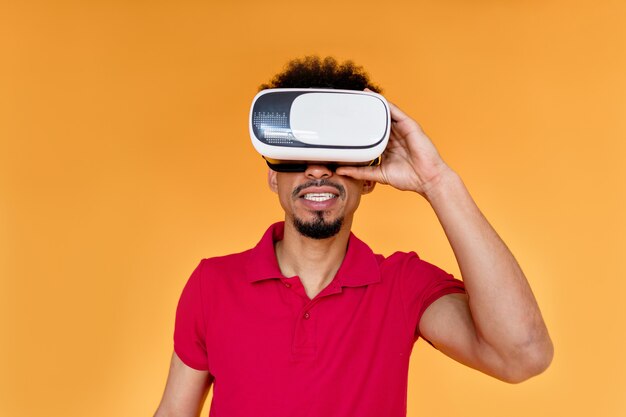 Joven afroamericano posando sobre una pared naranja con ropa de verano y casco de realidad virtual
