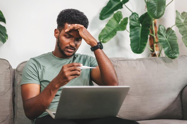 Joven afroamericano que trabaja en casa usando una computadora portátil para trabajar en la sala de estar tratamiento de dolor de cabeza cita en línea para una consulta médica