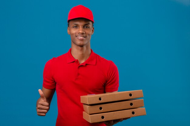 Joven afroamericano repartidor vistiendo camisa polo roja y gorra con pila de cajas de pizza mostrando los pulgares y sonriendo sobre azul aislado