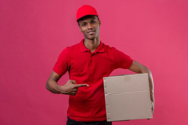 Joven afroamericano repartidor vistiendo polo rojo y gorra con caja de cartón y apuntando a la caja con el dedo sonriendo sobre rosa aislado