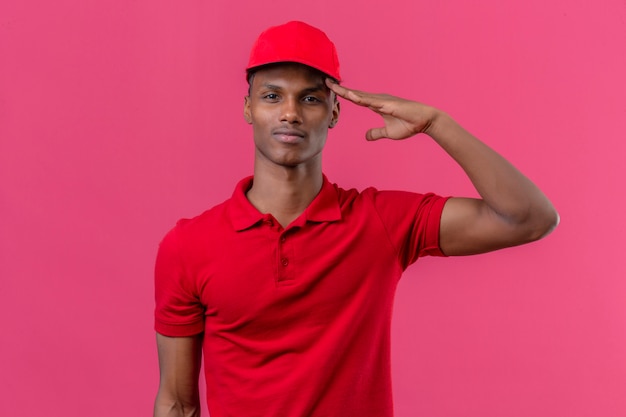 Joven afroamericano repartidor vistiendo polo rojo y gorra con cara seria saludando sobre rosa aislado