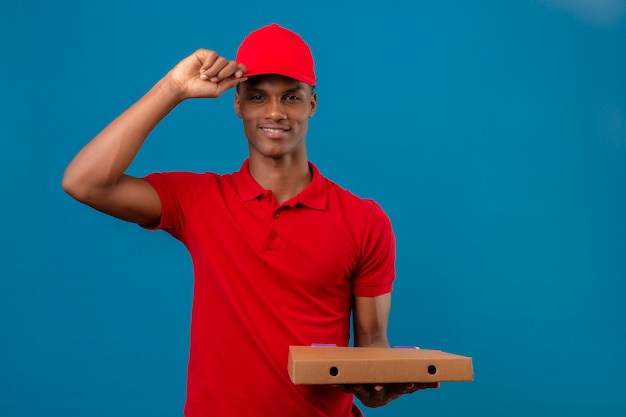 Joven afroamericano repartidor vistiendo polo rojo y gorra haciendo gesto de saludo tocando su gorra sobre azul aislado