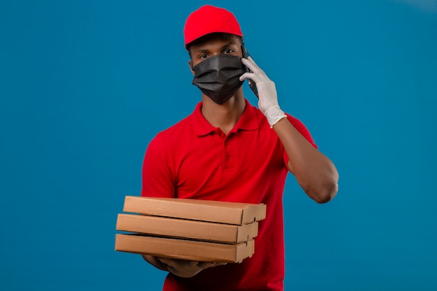 Joven afroamericano repartidor vistiendo polo rojo y gorra en máscara protectora y guantes con pila de cajas de pizza mientras habla por teléfono inteligente sobre azul aislado