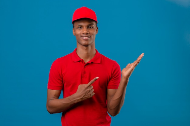 Joven afroamericano repartidor vistiendo polo rojo y gorra mostrando y apuntando hacia el lado mirando a la cámara con una sonrisa en la cara sobre azul aislado
