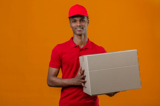 Joven afroamericano repartidor vistiendo polo rojo y gorra con paquete caja con sonrisa en la cara sobre naranja aislada