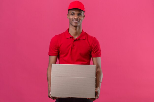 Joven afroamericano repartidor vistiendo polo rojo y gorra de pie con caja de cartón sonriendo sobre rosa aislado