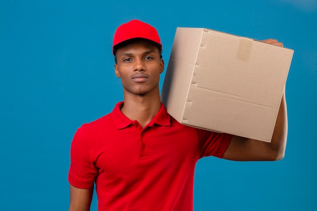 Joven afroamericano repartidor vistiendo polo rojo y gorra de pie con la caja en el hombro seguro mirando más azul aislado