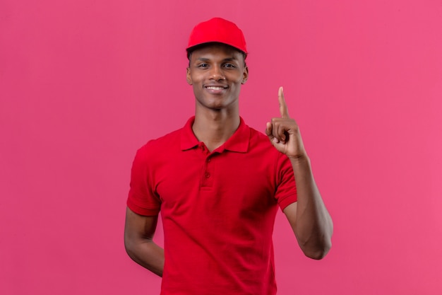 Joven afroamericano repartidor vistiendo polo rojo y gorra de pie con el dedo sonriendo sobre rosa aislado