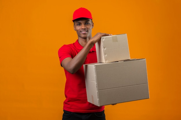 Joven afroamericano repartidor vistiendo polo rojo y gorra sosteniendo cajas con gran sonrisa sobre naranja aislada