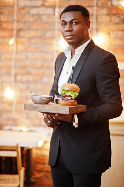Un joven afroamericano respetable con traje negro sostiene una bandeja con una hamburguesa doble contra la pared de ladrillos del restaurante con luces