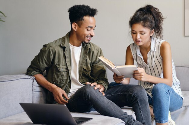 Joven afroamericano sonriente y bonita mujer asiática leyendo soñando un libro juntos en un moderno espacio de trabajo conjunto