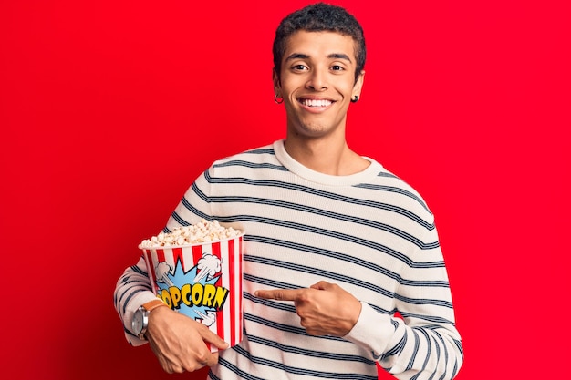 Joven afroamericano sosteniendo palomitas de maíz sonriendo feliz señalando con la mano y el dedo