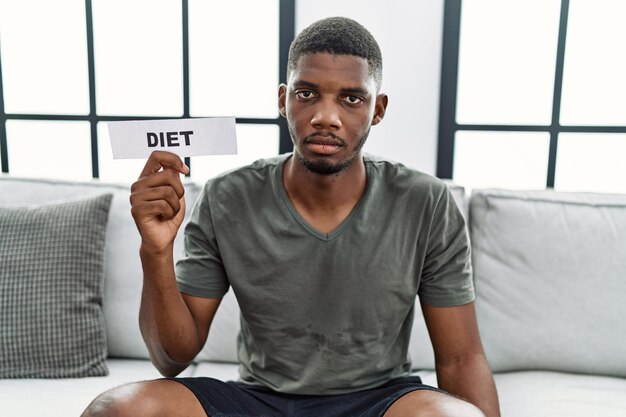 Joven afroamericano sosteniendo pancarta de dieta sentado en el sofá en casa pensando en actitud y expresión sobria que parece seguro de sí mismo