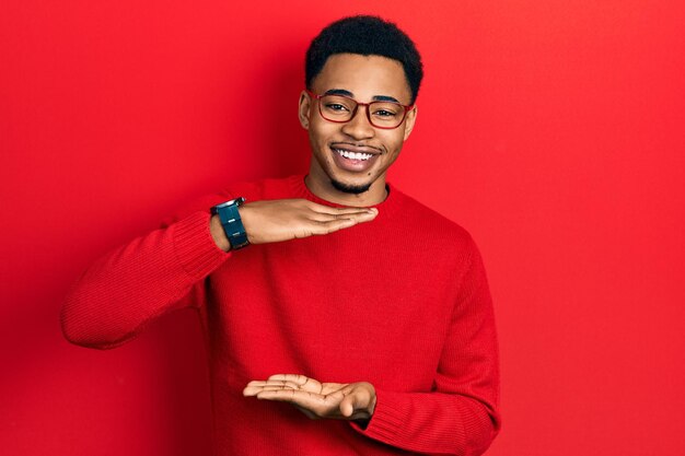 Joven afroamericano vestido con ropa informal y gafas gesticulando con las manos mostrando un símbolo de medida de signo grande y grande sonriendo mirando el concepto de medición de la cámara
