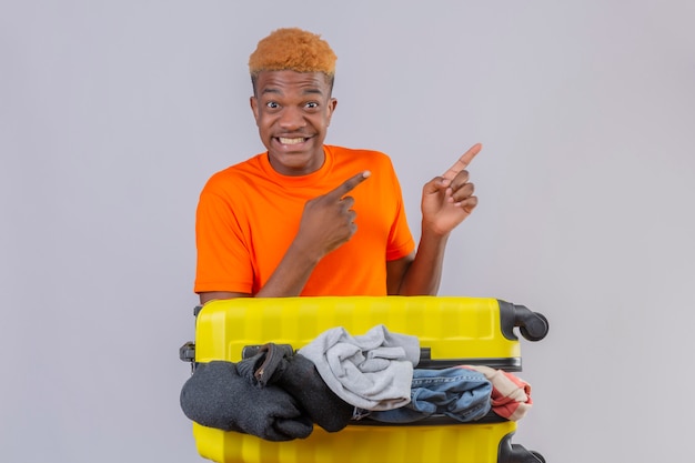 Joven afroamericano vistiendo camiseta naranja con maleta de viaje llena de ropa mirando a cámara sonriendo optimista y alegre apuntando con los dedos hacia un lado sobre fondo blanco.