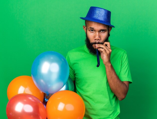 Joven afroamericano vistiendo gorro de fiesta soplando silbato de fiesta aislado en la pared verde