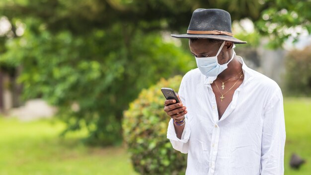 Joven afroamericano vistiendo una mascarilla protectora usando su teléfono al aire libre