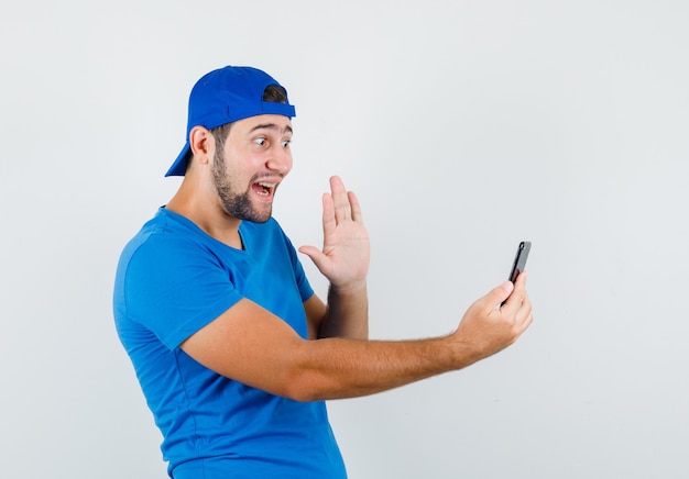 Joven agitando la mano en el chat de video en camiseta azul y gorra y mirando alegre