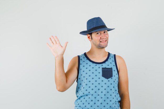 Joven agitando la mano para saludar en camiseta azul, sombrero y mirando jovial. vista frontal.