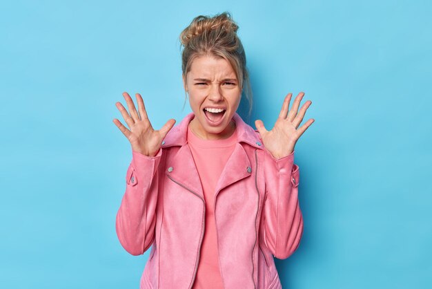 Una joven agresiva y descontenta levanta las palmas de las manos grita furiosamente expresa emociones negativas mantiene la boca abierta usa poses de chaqueta rosa contra el fondo azul del estudio. no puedo soportar esto mas